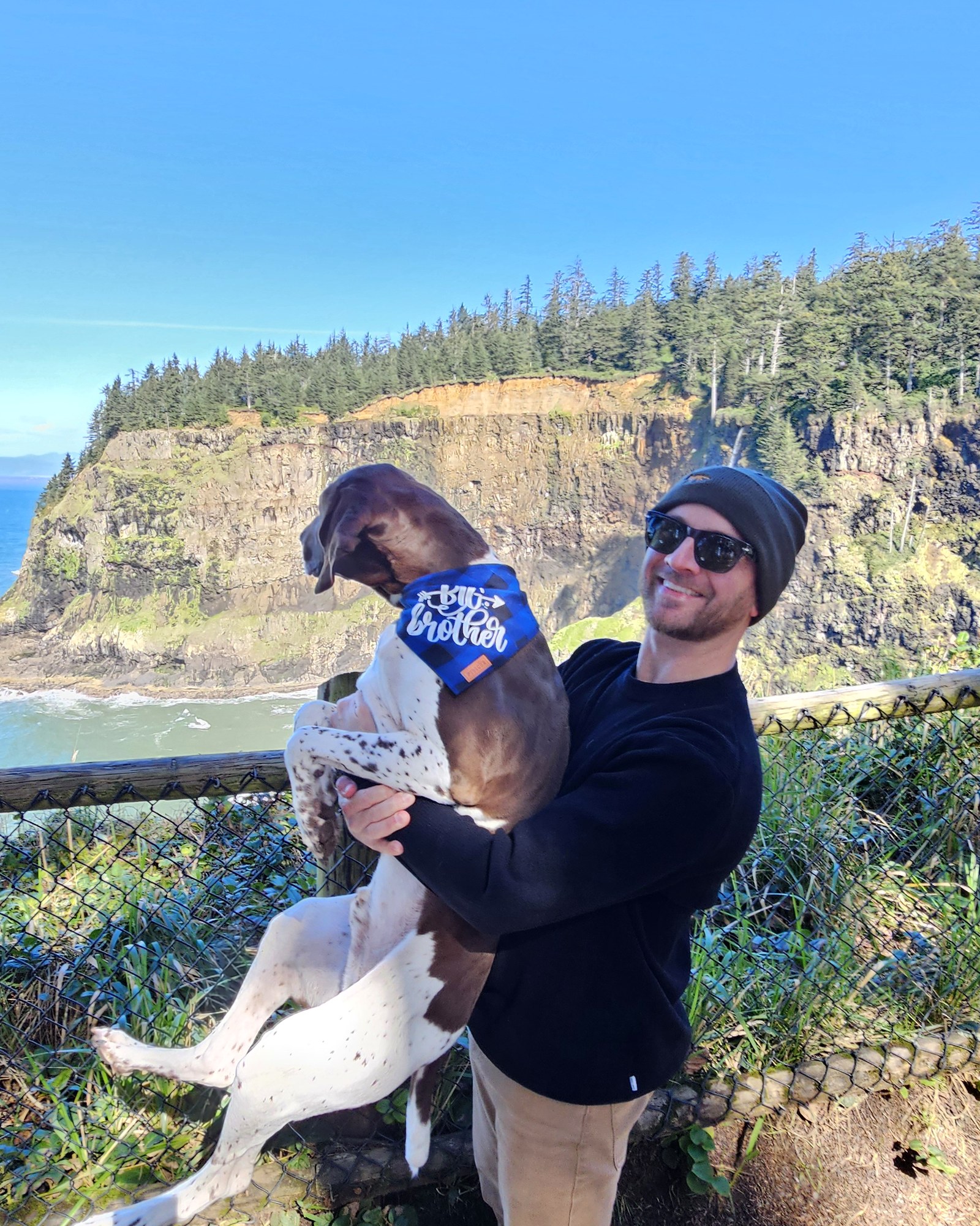 John holding Mac, his German Shorthaired Pointer, who is wearing a blue 'big brother' bandana, on a bluff at Cape Meares overlooking the Pacific